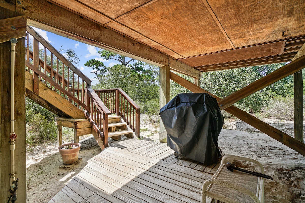 A shaded outdoor space features wooden decking, leading to a set of stairs and a covered area. A barbecue grill is covered nearby, with a potted plant and a white chair positioned on the wood flooring, offering a relaxed atmosphere in the surrounding nature.