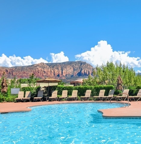 An outdoor heated swimming pool is surrounded by tan lounge chairs, offering views of the red rock formations in the distance. Trees and landscaping frame the area, while a bright blue sky with fluffy white clouds stretches overhead.