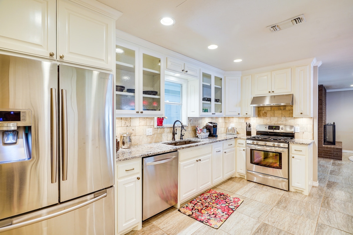 A well-equipped kitchen features stainless steel appliances, including a refrigerator and oven. Cream-colored cabinetry offers ample storage, with glass-fronted shelves displaying dishware. A granite countertop runs along the preparation area, complemented by a decorative rug and soft lighting throughout the space.
