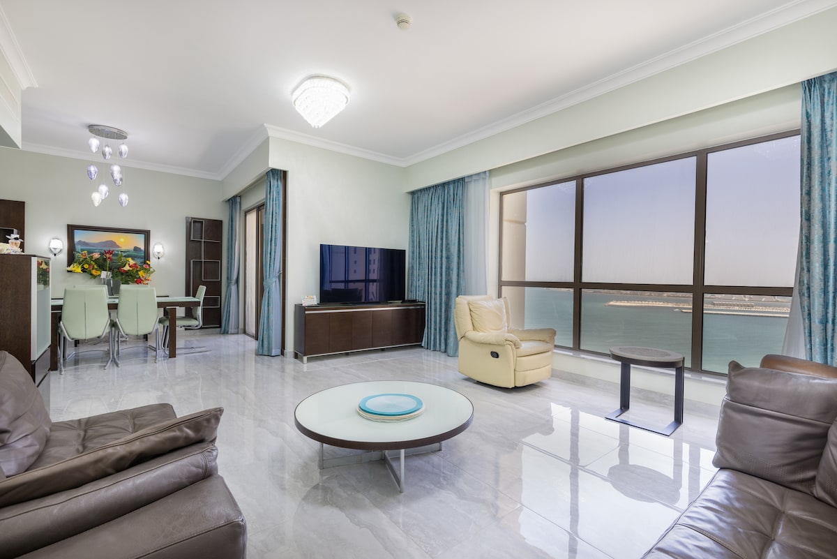 A spacious living area featuring large floor-to-ceiling windows that provide a direct view of the sea. Elegant furniture includes a light leather armchair and a contemporary coffee table set on a polished marble floor. The dining area is positioned nearby, with a modern table and seating.