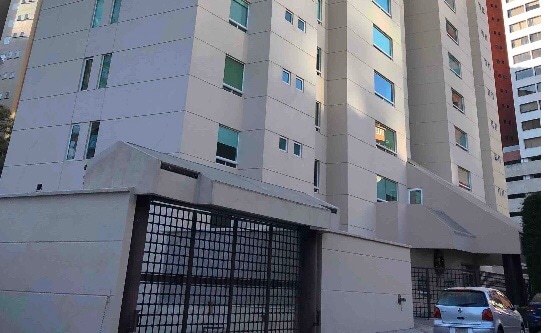 The building exterior features a modern gray facade with symmetrical windows providing natural light. A gated entrance is visible, indicating security, alongside a parking space for vehicles. The setting is surrounded by urban structures, highlighting the proximity to city amenities.