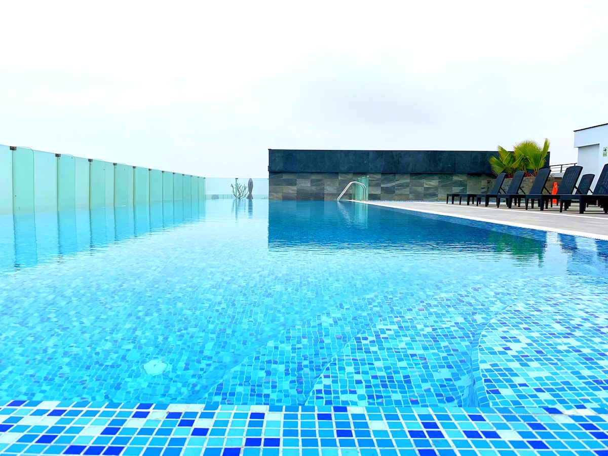 An expansive rooftop infinity pool extends into the horizon, featuring vibrant blue mosaic tiles. Loungers are arranged on the surrounding deck, offering views over the water. A clear barrier provides safety while maintaining unobstructed visibility of the landscape.