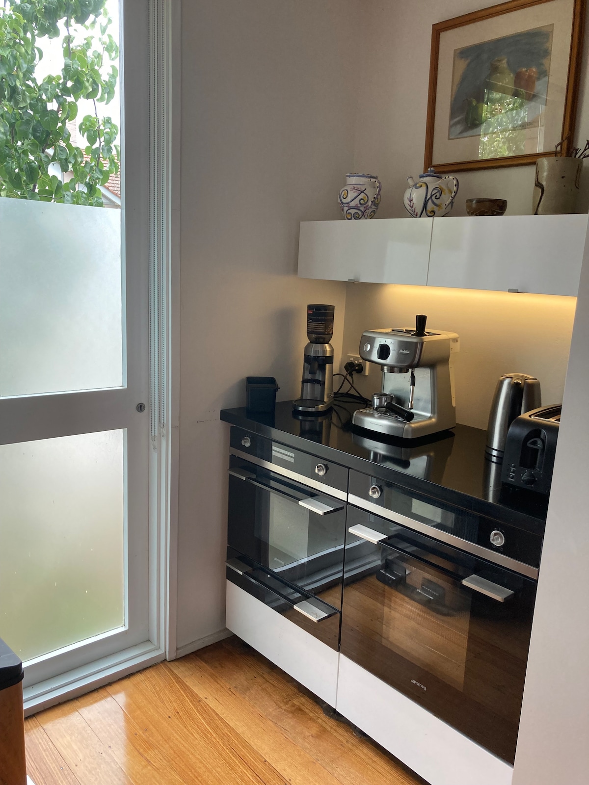 A modern kitchen corner is equipped with sleek black cabinetry and stainless steel appliances. A coffee maker and kettle are positioned on the countertop, while natural light filters through a nearby frosted glass window. Decorative items are displayed on the shelves above.