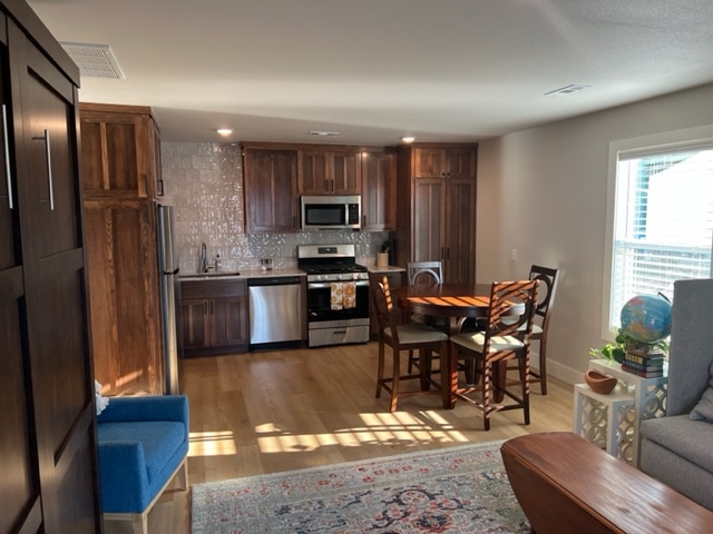A modern kitchen is visible, featuring stainless steel appliances and dark wood cabinetry. A dining table with four chairs is positioned nearby. Sunlight brightens the space, highlighting the wooden flooring and a comfortable seating area with a blue chair and a gray couch.