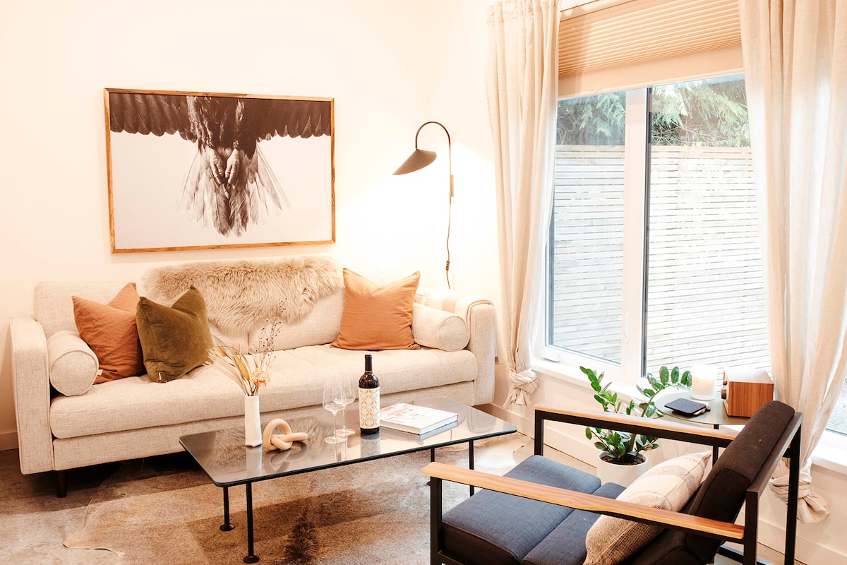 A cozy living area features a light-colored sofa adorned with decorative pillows and a soft throw. A glass coffee table is positioned in front, with a small plant nearby. Natural light enters through large windows, complemented by sheer curtains and a contemporary floor lamp.