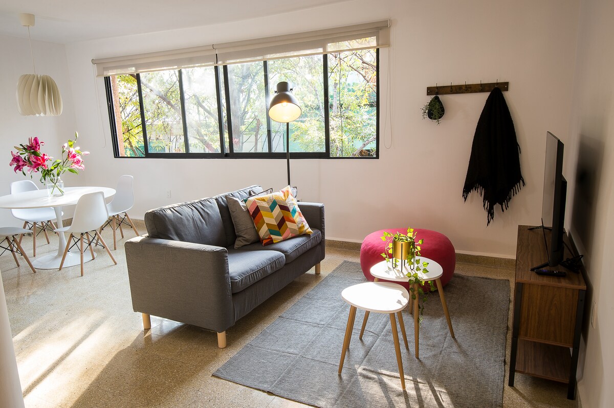An airy living space features large windows allowing natural light to fill the room. A cozy gray sofa adorned with colorful cushions faces two small tables and a television. In the background, a round dining table with white chairs is set beside a plant. A soft rug adds comfort.