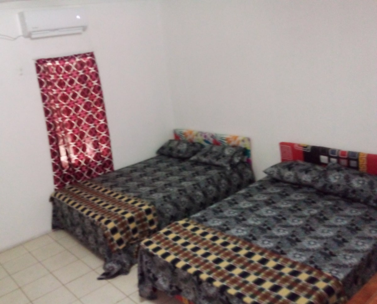 A bright sleeping area is furnished with two spacious beds, each dressed in patterned covers. A window with bold, colorful curtains allows natural light to enter the room, which features neutral walls and tiled flooring.