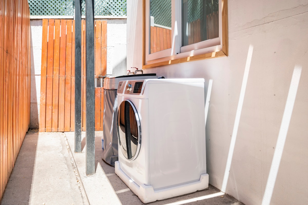 A dedicated laundry area features a washing machine and dryer positioned beside a wall. Natural light enters through a nearby window, illuminating the appliances and a concrete floor. The space is enclosed by wooden fencing, providing a sense of privacy.