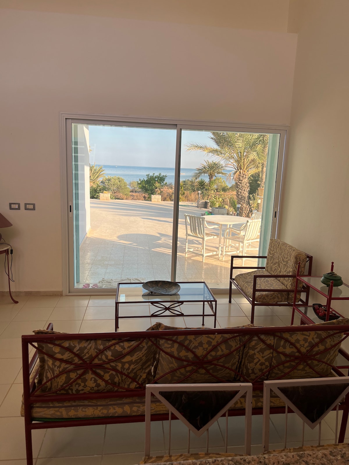 A comfortable seating area is displayed, featuring two armchairs and a glass-top table. A large sliding door opens to an outdoor patio with views of palm trees and the sea, allowing natural light to fill the space.