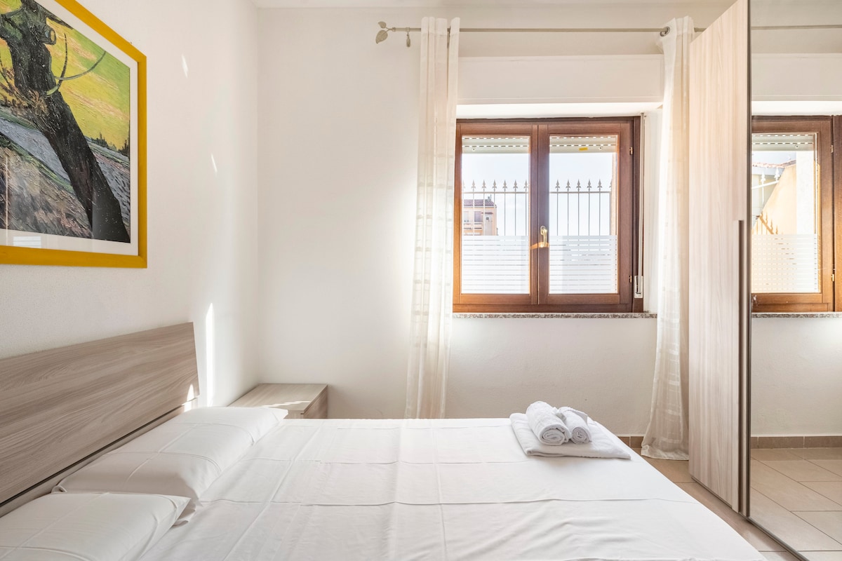 A bright sleeping area is highlighted by natural light streaming from two windows with sheer curtains. A spacious bed is complemented by simple bedside tables, while neatly rolled towels are arranged at the foot of the bed. A piece of artwork adds a touch of color to the wall.
