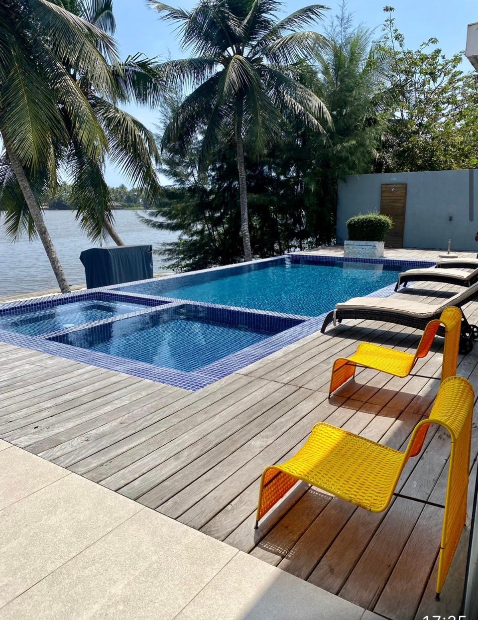 A serene outdoor area is depicted, featuring a tranquil swimming pool bordered by a wooden deck. Two lounge chairs in vibrant yellow are positioned for relaxation, and palm trees provide a lush backdrop. A jacuzzi is integrated into the pool area, enhancing the space's appeal.