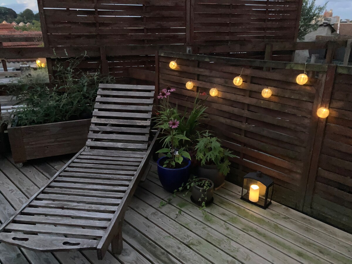A wooden lounger is placed on a deck, surrounded by potted plants and soft lighting. Lanterns hang along a wooden privacy screen, creating a warm ambiance alongside a solitary candle on the floor. The setting offers a peaceful space for relaxation.