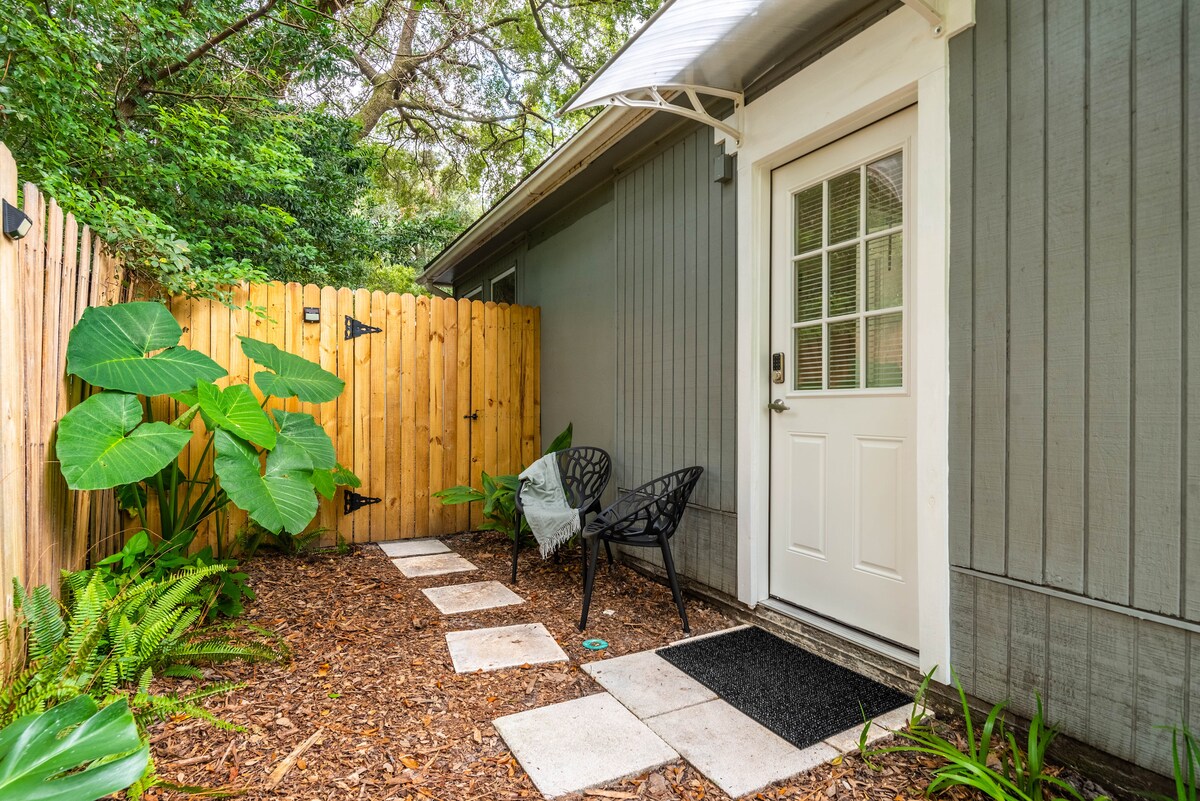 A private entrance is visible, featuring a light-colored door surrounded by greenery. The pathway leading to the door is made of square stones, bordered by landscaped areas with large leaves and mulch. Two black chairs are placed near the entrance, adding to the welcoming outdoor space.
