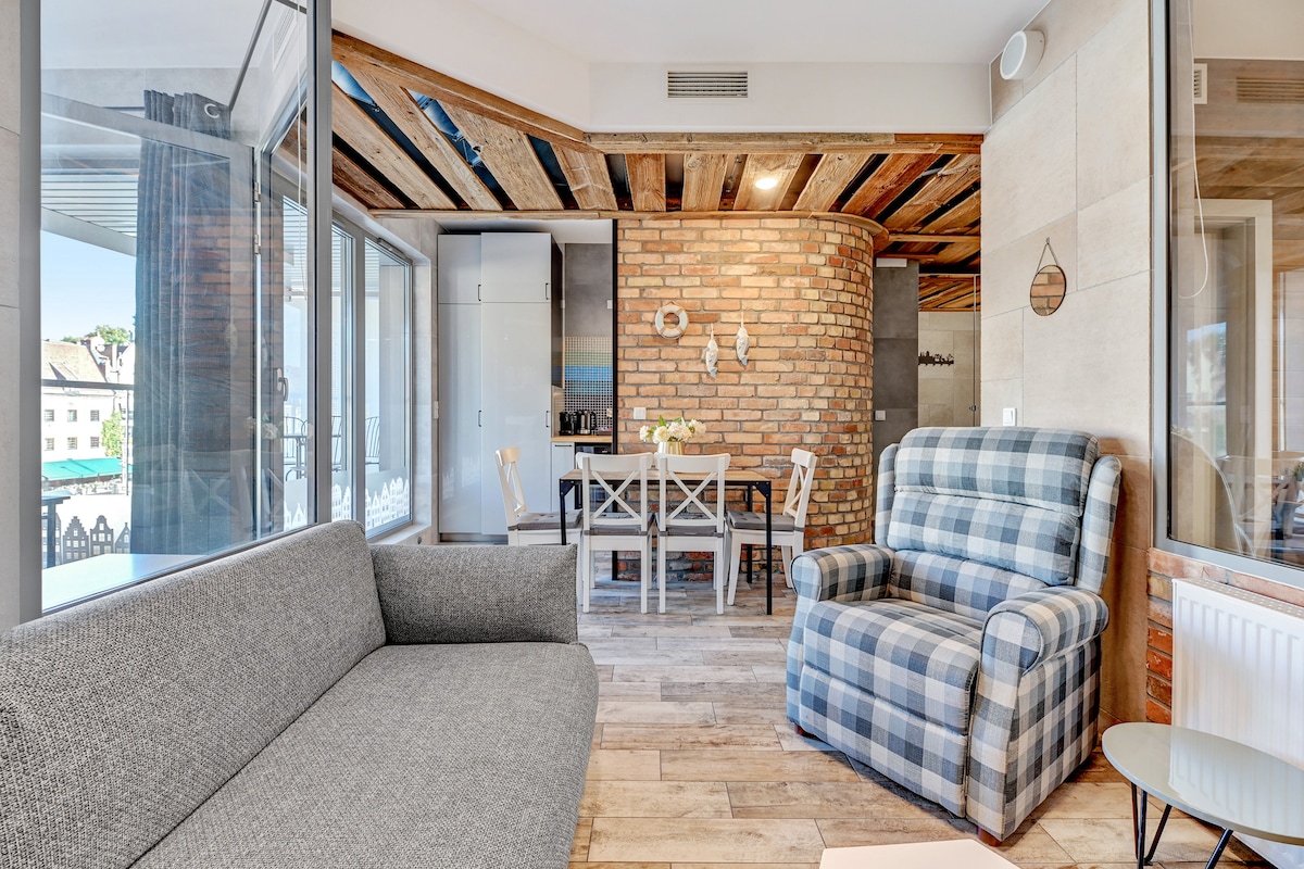 A bright living area is presented with wooden beams and an open layout. A gray sofa and a checkered armchair are positioned for comfort. A dining table with chairs sits nearby, while a kitchenette is partially visible in the background, creating a functional and welcoming space.