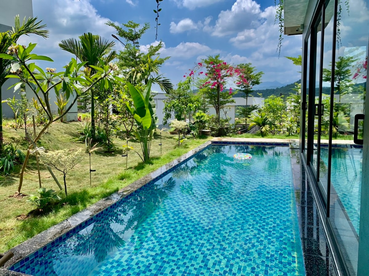 A private pool is surrounded by lush greenery and tropical plants, reflecting the clear sky above. The pool features vibrant blue mosaic tiles and is bordered by well-maintained grass, creating an inviting outdoor space.