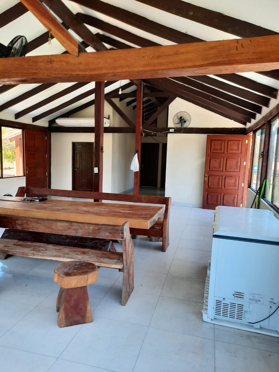 A spacious dining area is highlighted by a large wooden table and matching benches, complemented by a uniquely shaped wooden stool. High wooden beams create an airy ambience, while an air conditioning unit is mounted on the ceiling, providing comfort. The room is well-lit by natural light from nearby windows.