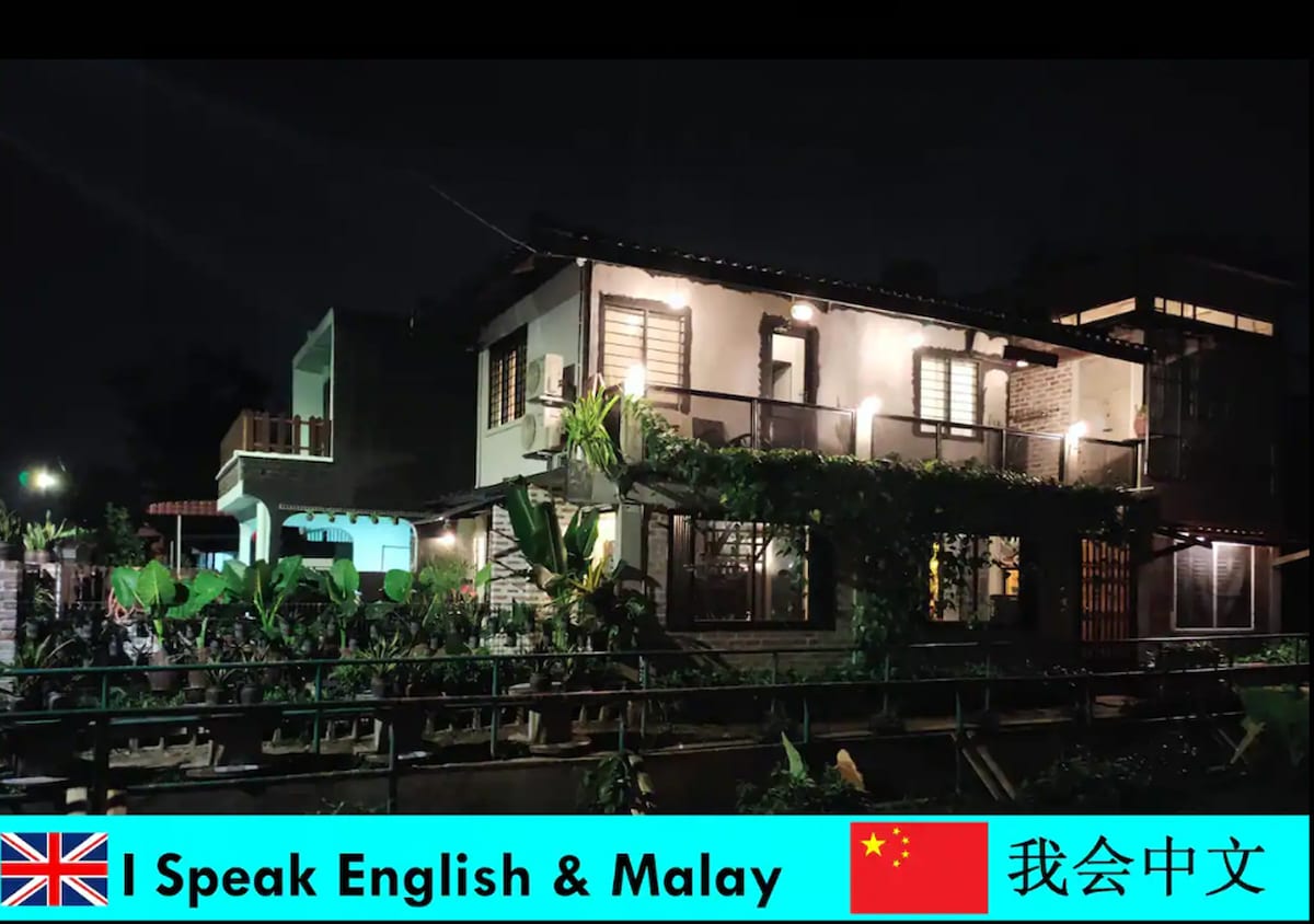 The exterior of a two-story building is shown at night, featuring warm light spilling from windows. Lush greenery surrounds the home, and a balcony extends on the upper level, providing a welcoming ambiance. The structure blends traditional and modern architectural elements harmoniously.