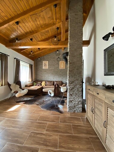 A cozy living area features a stone wall and wooden beams above. A comfortable seating arrangement includes soft chairs and a sectional couch. Natural light filters through the window, illuminating the space, while decorative elements like wall art and a cowhide rug add texture.