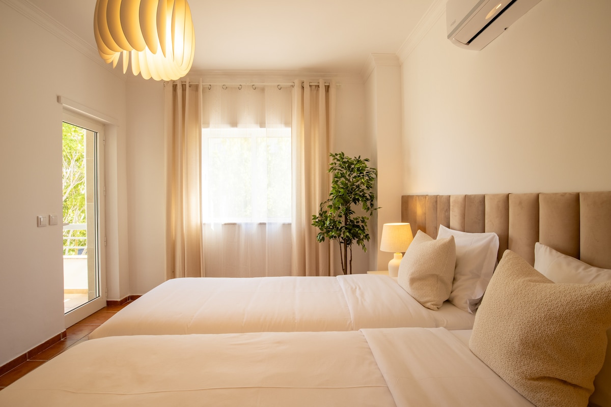 The twin bedroom features two individual beds positioned near a window, allowing natural light to brighten the space. A small bedside lamp and decorative plant add subtle touches. Soft curtains offer privacy and a sense of calm.