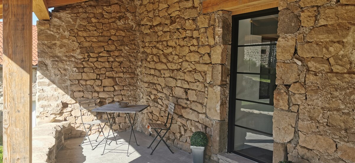 An outdoor seating area features a small table accompanied by two chairs, set against a rustic stone wall. A potted plant adds a touch of greenery, while large glass doors provide a view into the interior space.