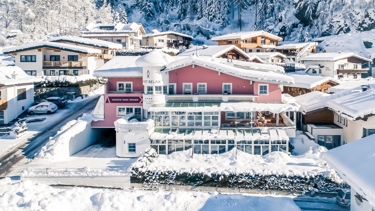 The building is surrounded by a snowy landscape, showcasing a modern design with multiple levels. Glass elements are visible, indicating spacious indoor areas. Nearby, traditional alpine-style buildings complement the scene, with snow covering the ground and rooftops, creating a serene winter atmosphere.
