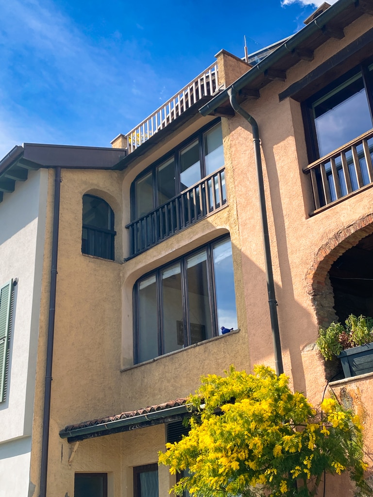 The exterior of a three-story house is shown, featuring a textured facade in warm tones and large windows that provide ample natural light. A balcony extends from the upper level, surrounded by plants. Lush greenery is visible at the base, adding to the inviting appearance.