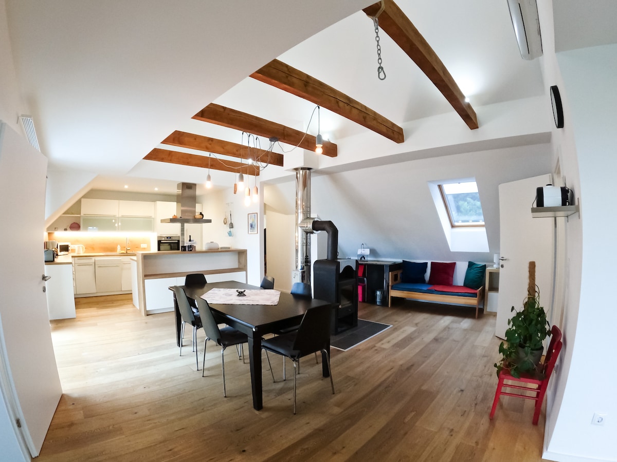 An open-plan living area features wooden beams overhead and large windows allowing natural light. A dining table is surrounded by black chairs, while a cozy seating area is visible nearby. A modern kitchen is equipped with essential appliances, enhancing the functional space.