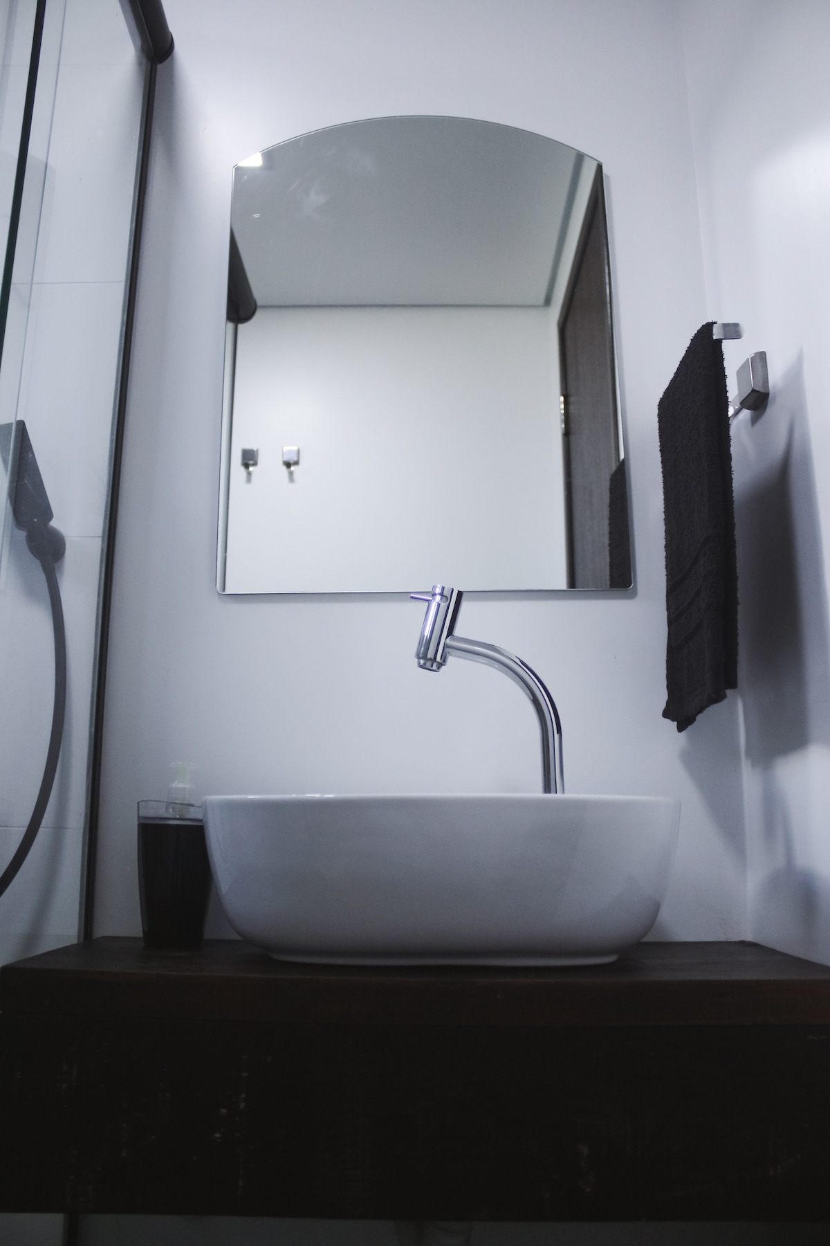 A modern bathroom features a sleek, white vessel sink atop a wooden countertop. Above the sink, a curved mirror reflects the light. A hand towel hangs on the side, and a soap dispenser is positioned on the countertop, creating a clean and functional space.