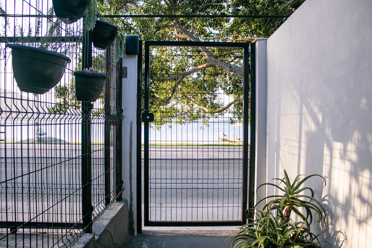 A secure gate is set within a fence, framed by trees and climbing plants. The gate opens onto a tranquil view of the waterfront across the street, allowing for easy access to the nearby lagoon.