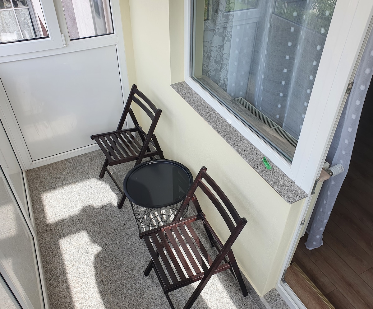 A small balcony area is presented, featuring two wooden chairs and a round, black table. Light floods the space, highlighting the tan floor and textured walls. A window with sheer curtains offers a view of the outside while maintaining a sense of privacy.