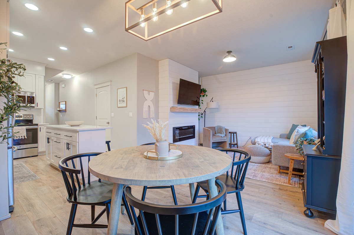A dining area is highlighted by a round wooden table surrounded by black chairs. A cozy living space is visible in the background, featuring a fireplace and a mounted television. Natural light fills the open layout, enhancing the modern, welcoming ambiance.