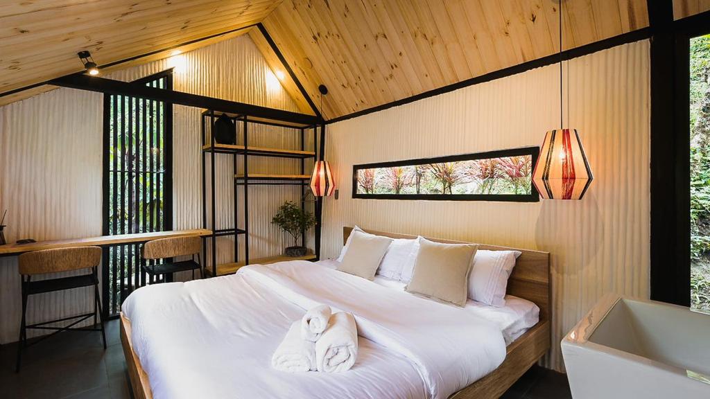 A well-appointed sleeping area features a spacious bed with soft linens and decorative pillows. Natural wood paneling creates a warm ambiance, while large windows offer views of greenery. Two pendant lights add a gentle glow, and a small workspace is included in the corner.