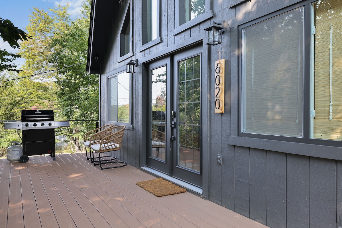 The front deck is shown with a seating area featuring a chair and a grill. The entrance displays double glass doors and a vertical wooden sign with the number '6020'. Sunlight filters through the surrounding trees.