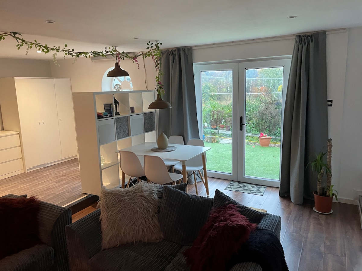 A modern living space features a dining area with a round table and white chairs, situated next to large glass doors that open to a garden. Soft furnishings are arranged on a couch, with decorative plants and shelving units visible in the background.