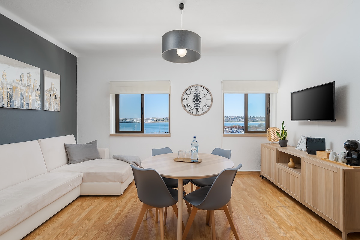A light-filled living area features a comfortable corner sofa and a round dining table surrounded by gray chairs. Two windows offer views of the river and nearby scenery. A modern wall clock and a television are visible, complementing the warm wooden flooring.