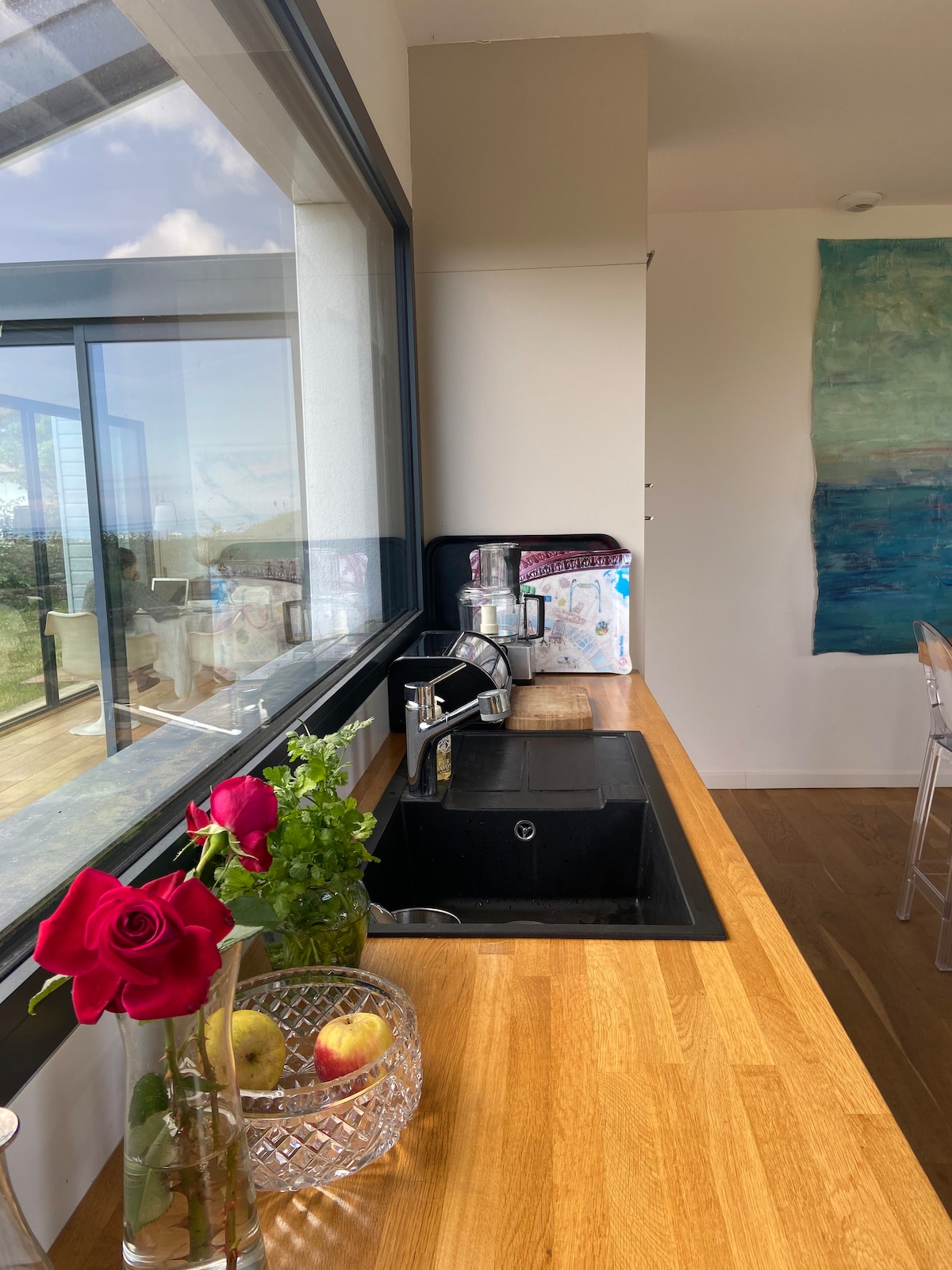 A modern kitchen countertop is presented with a black sink, adorned by a crystal vase containing red roses and green foliage. Fresh apples rest nearby, and the space is illuminated by natural light from large windows showcasing scenic outdoor views.