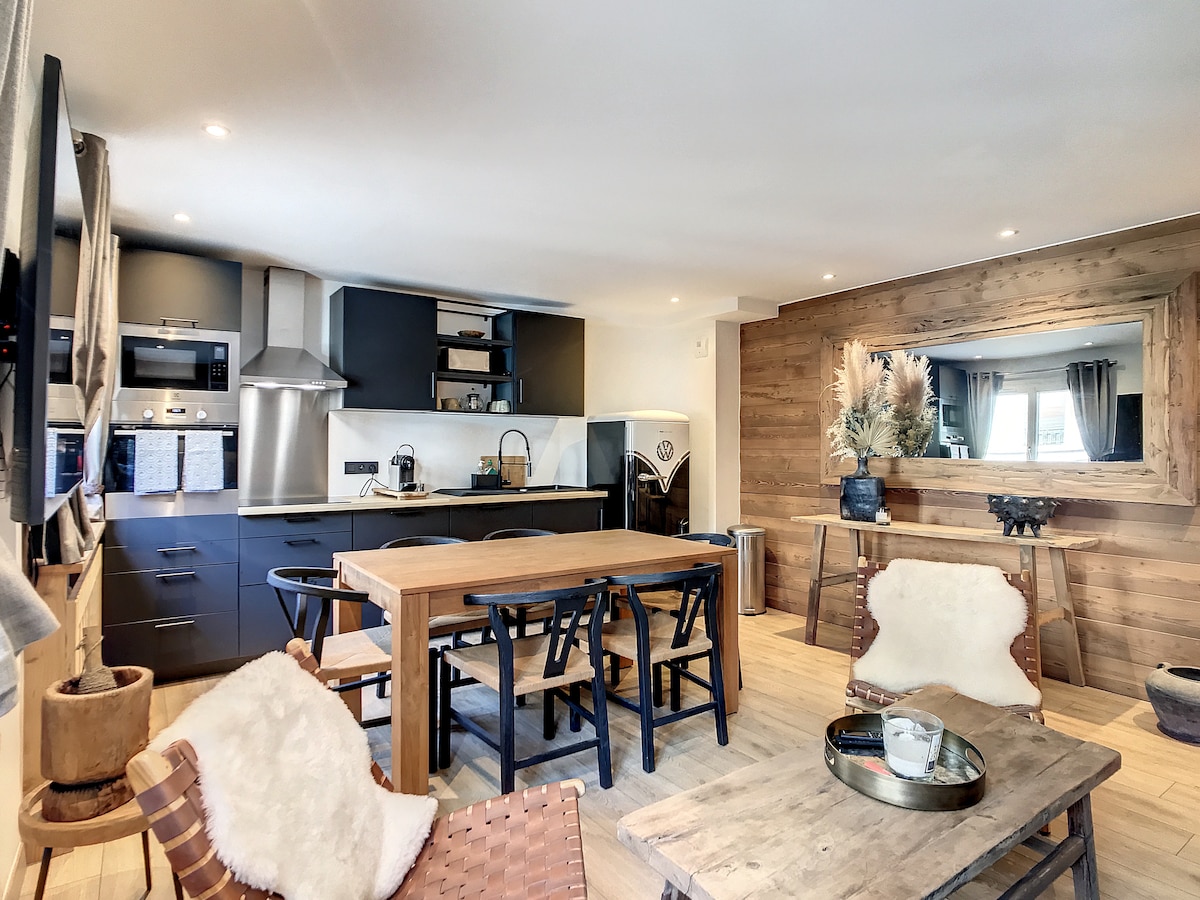 A modern kitchen area is complemented by a spacious dining table surrounded by chairs. Wood accents enhance the cozy aesthetic. The kitchen features sleek cabinetry, an integrated oven, and essential appliances, while natural light streams in through nearby windows.