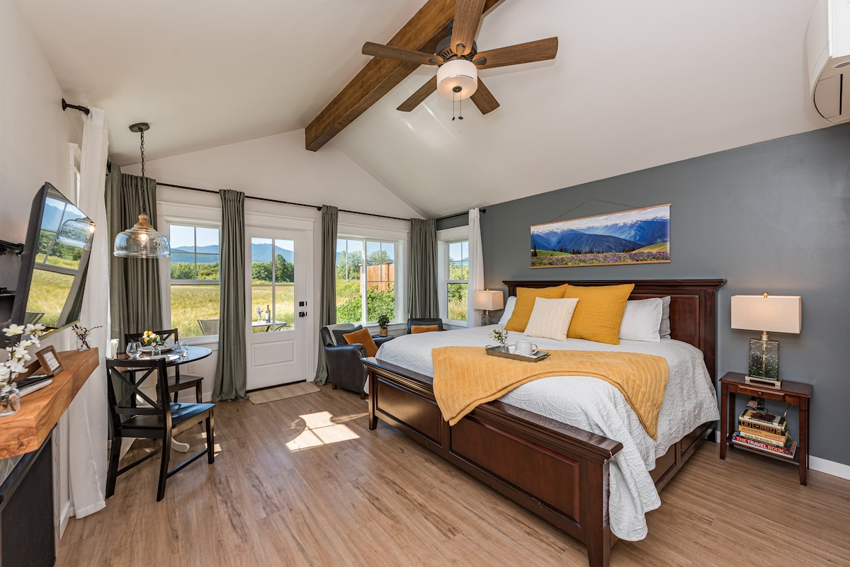 A spacious bedroom features a king bed with soft linens and a mustard yellow throw blanket. Large windows showcase expansive mountain views. A small dining table with two chairs sits in a corner. Warm wood beams accentuate the high ceiling, enhancing the cozy atmosphere.