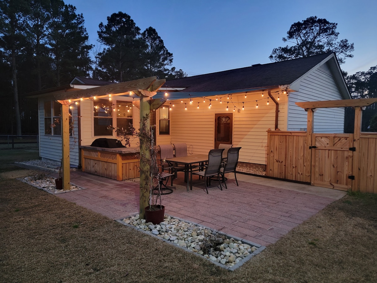 A covered outdoor seating area features a spacious table surrounded by multiple chairs. Soft patio lights illuminate the space, creating a welcoming environment. The yard is graced with natural stone accents and fenced for added privacy.