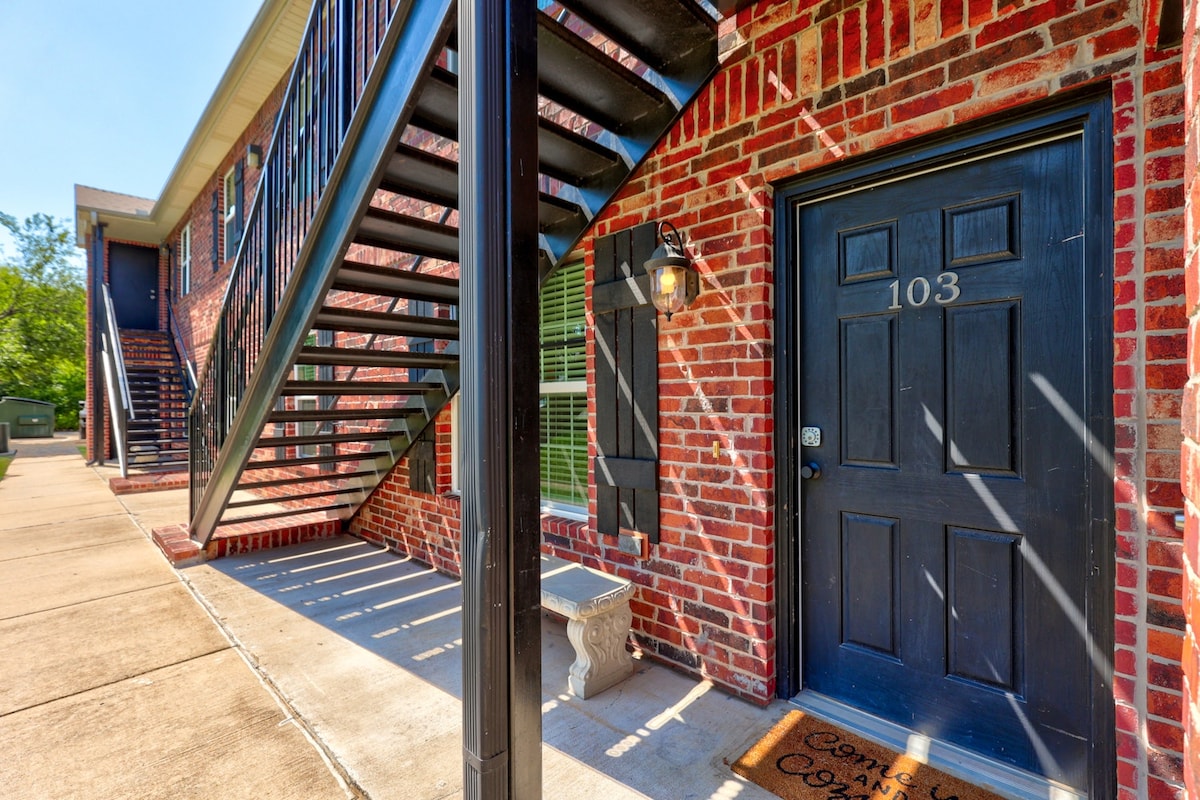 The exterior features a brick facade with a well-maintained entrance. The door, marked 103, is flanked by a stone bench and a welcome mat. A black iron staircase leads to the upper units, with sunlight casting defined shadows on the concrete walkway.