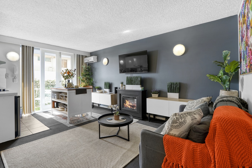 A modern living area features a comfortable couch with an orange throw and decorative pillows. A flat-screen TV is mounted on the wall, complemented by a small coffee table in front. A console table holds decorative plants. Natural light streams through large windows.