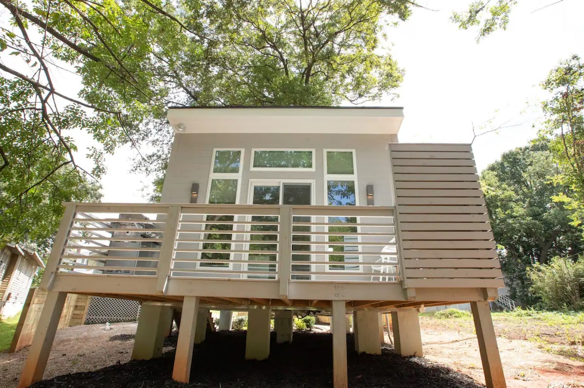 A compact modern tiny home is elevated on stilts, featuring a welcoming front deck surrounded by a railing. Large windows are visible, allowing natural light to fill the interior. Lush greenery can be seen in the background, enhancing the serene environment.