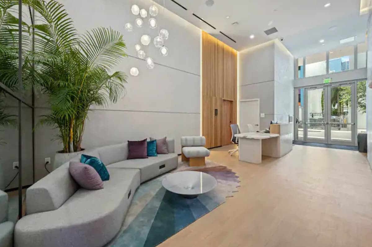 A contemporary lobby is featured, with soft seating arranged on a light wood floor. Soft textures and colors are presented through cushions in various shades on a curved gray sofa. An elegant chandelier hangs overhead, complemented by large glass doors leading outside.