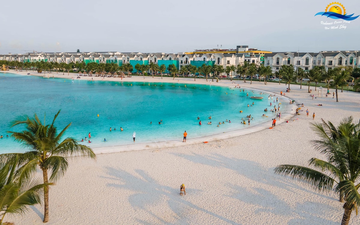A scenic beach setting is captured, featuring a curved shoreline with soft white sand. Turquoise waters gently lap the shore, while groups of people can be seen enjoying the beach. Palm trees line the edge, providing a natural frame to the tranquil scene.