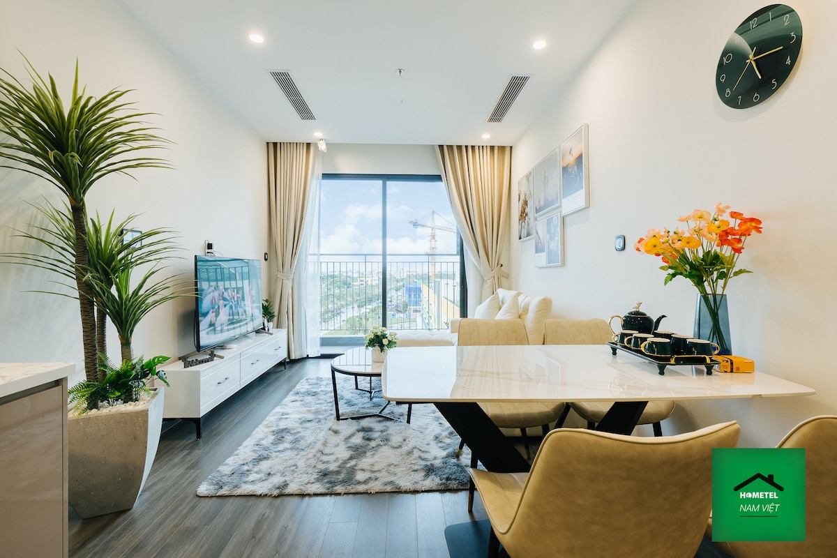 A bright living area is highlighted by large windows that provide city views. A cozy sofa and a marble dining table are complemented by soft decor and greenery. A wall-mounted TV and modern clock add functional touches to the elegantly styled space.