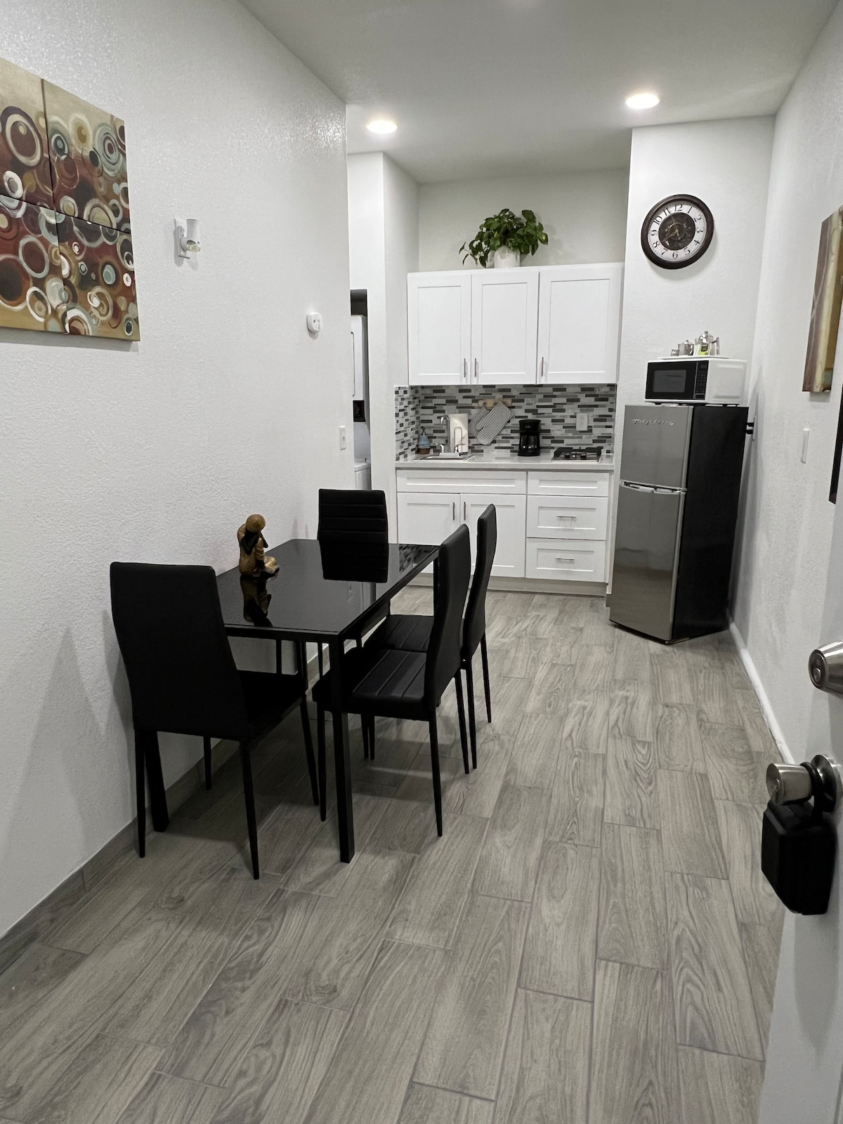 A modern dining area features a black glass table surrounded by four sleek chairs. A small decorative figure is placed on the table. The kitchen area is visible in the background, showcasing white cabinetry and a stainless-steel refrigerator.