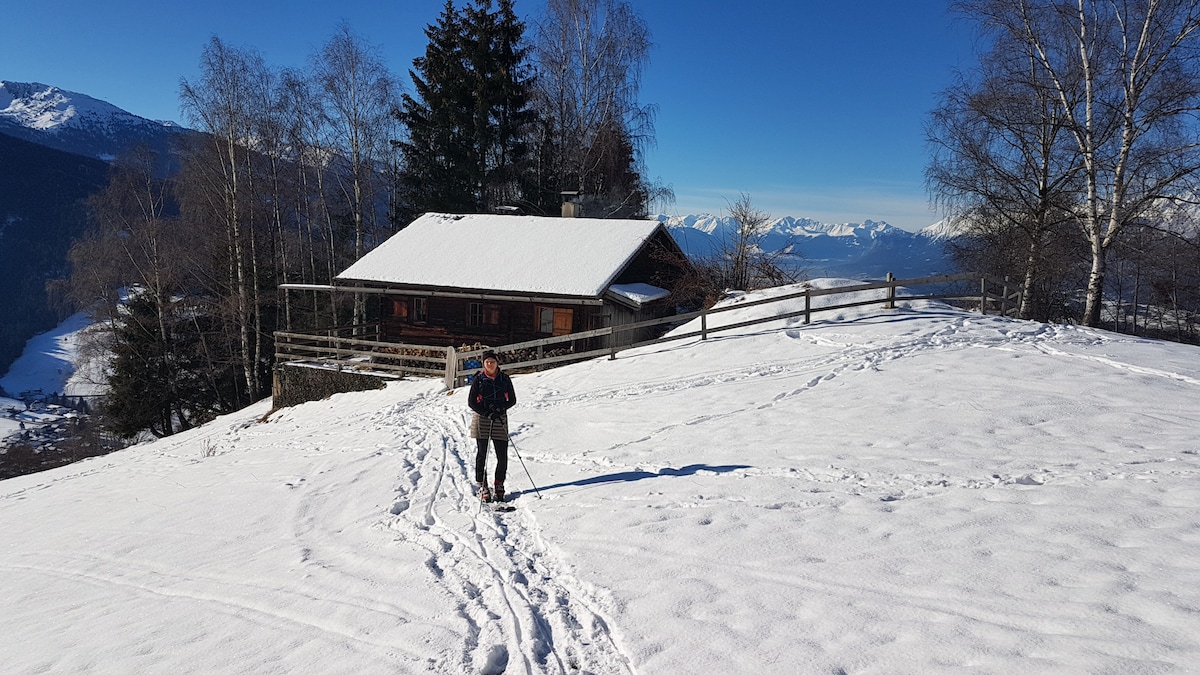 A snowy landscape is shown, featuring a rustic cabin surrounded by trees and mountains. A person is seen skiing along a path marked in the fresh snow, with the cabin's sloped roof covered in snow reflecting sunlight. The distant mountain range serves as a scenic backdrop.