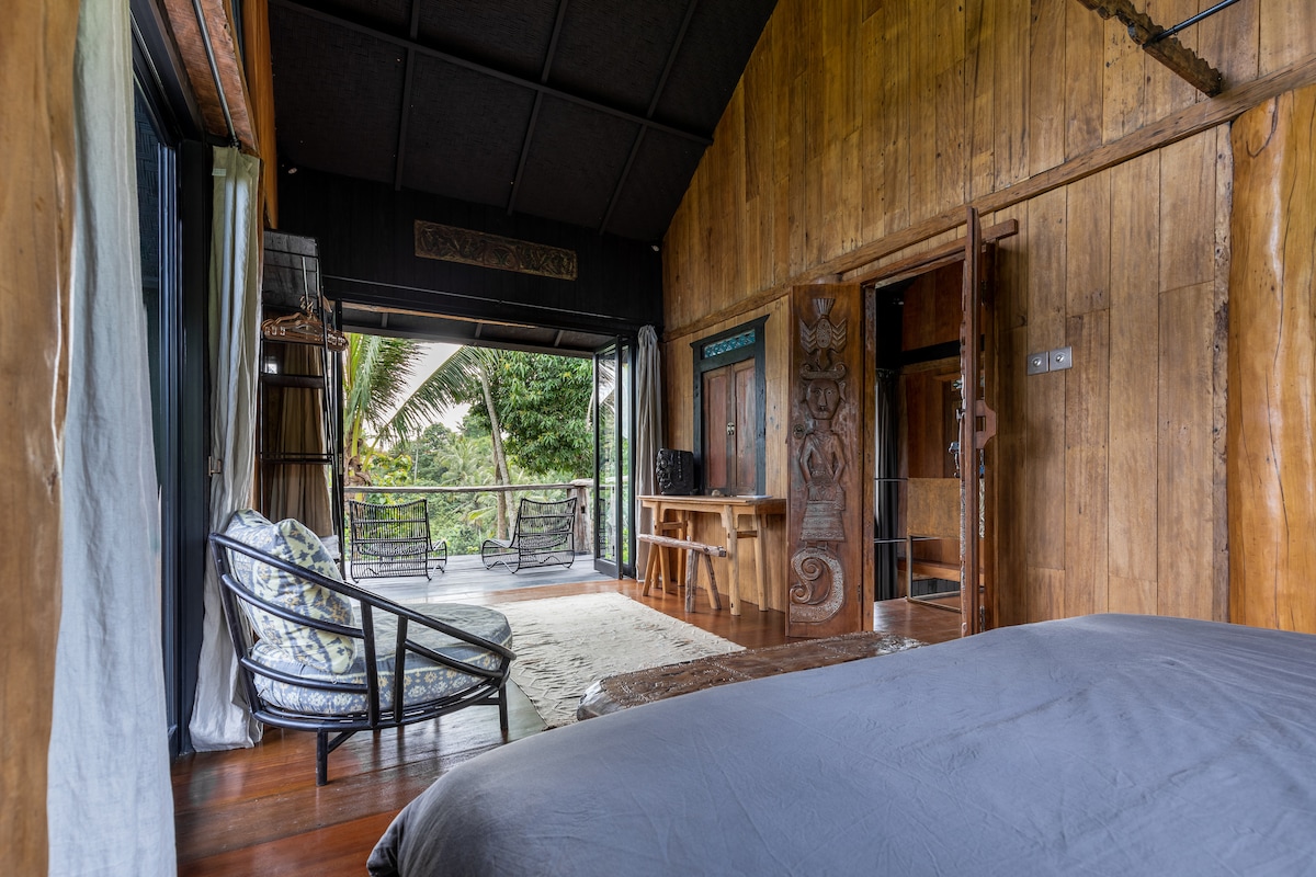A cozy bedroom showcases rich wooden walls and a spacious bed. A private balcony with outdoor seating overlooks lush greenery. Natural light floods the room through floor-to-ceiling glass doors, creating a serene atmosphere. A small workspace is also visible near the entrance.