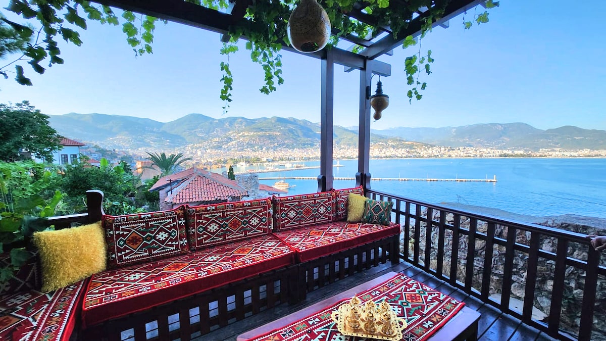 A seating area is situated on a wooden terrace, offering panoramic views of the coastline and mountains. Colorful, patterned cushions adorn the spacious bench, complemented by a decorative rug. Lush greenery surrounds the space, enhancing the serene atmosphere. Natural light filters through the vines overhead.