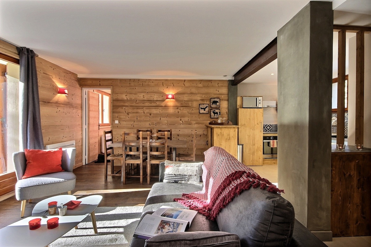 A spacious lounge area features a comfortable sectional sofa with a red throw blanket, a coffee table with decorative items, and textured rugs. Natural light enters through large windows, illuminating the wood-paneled walls. The adjoining dining space includes a wooden table and chairs.